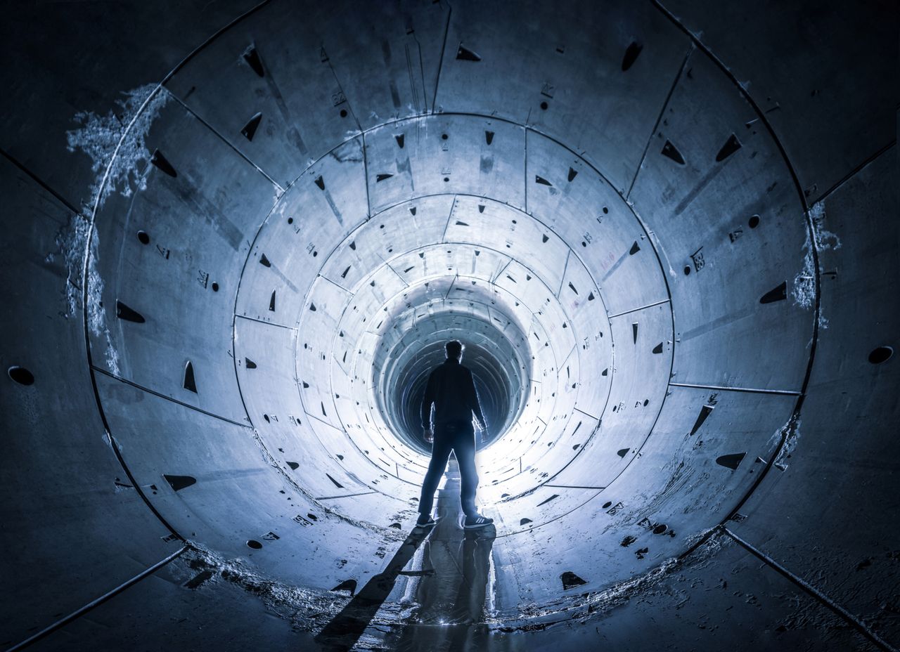A mysterious silhouette stands in the illuminated entrance of a concrete tunnel.