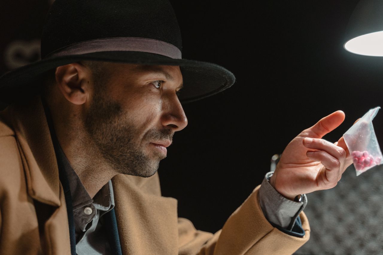 A man in a coat and hat holds a bag of red pills under a spotlight.